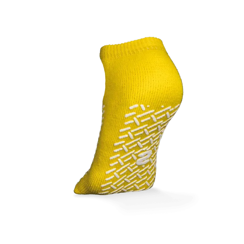 Ein einzelner gelber ECO-PLUS Anti-Rutsch Einwegsocken beidseitig beschichtet von AMPri Handelsgesellschaft mbH steht aufrecht auf weißem Hintergrund, mit weißen geometrischen Antirutsch-Griffen und einem zentralen Logo, ideal für medizinische Bereiche.