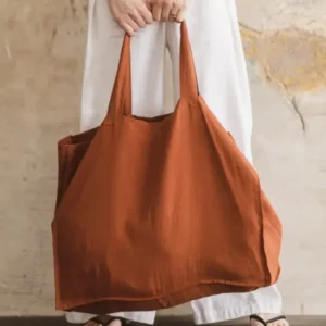 Woman holding large terracotta linen tote bag by both handles against stone wall background